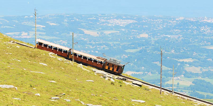 Wooden train heading down hill down a steep mountain in the foreground with the sea and town in the background.