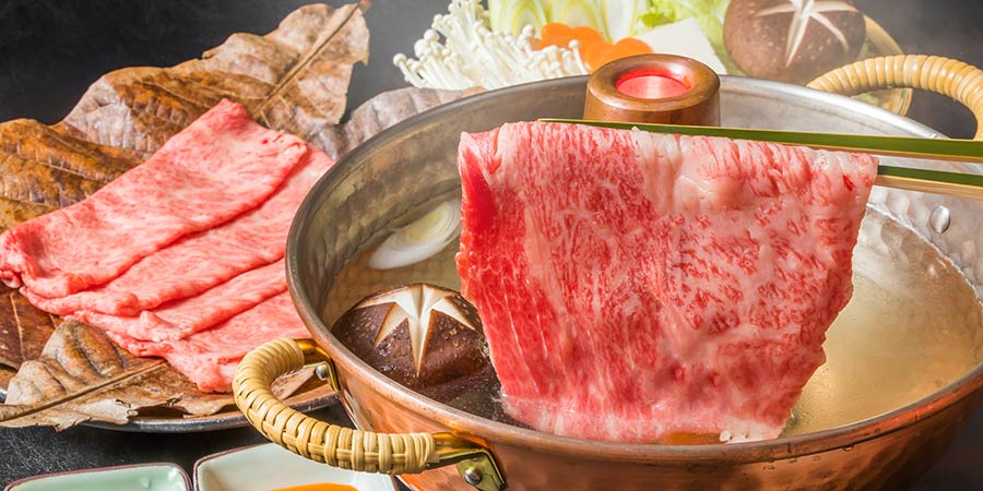 A meal of shabu-shabu, a piece of raw marbled beef is cooked in a pot of hot broth, surrounded by mushrooms and vegetables.