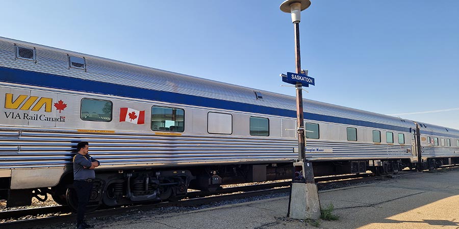 A modern silver VIA Rail train sits on a sunny platform on a clear blue day.