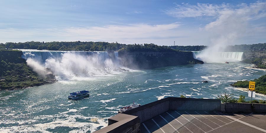 Water rushes over the Niagara Falls creating clouds of white spray and a small blue tourist boat sits in the middle of the water.