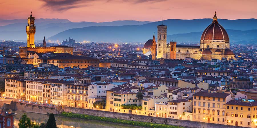 Panoramic view of Florence, Italy, highlighting the city’s iconic skyline with the Florence Cathedral (Duomo) and its red-tiled dome standing prominently. The Arno River flows through the city, in the foreground with the Tuscan hills in the background under a soft, golden sunset."