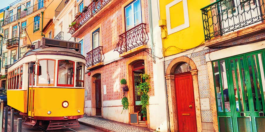 A bright yellow tram travels down the track of a Lisbon street with brightly coloured doors.