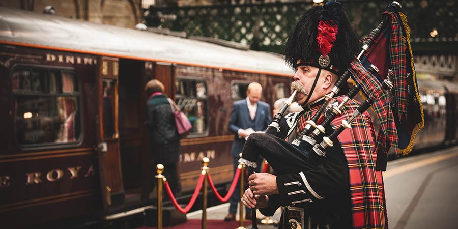 A man dressed in traditional Scottish military unform plays the bagpipes while passengers board the Royal Scotsman train.