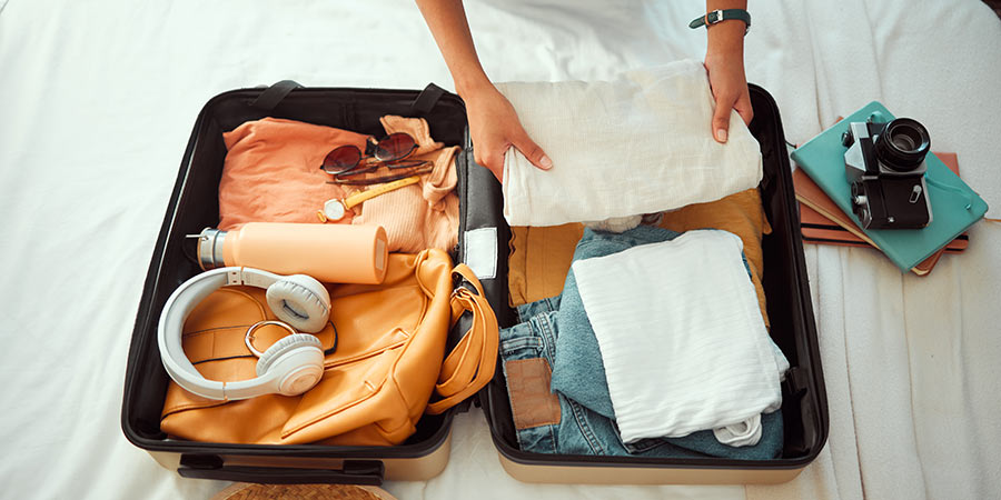 A person is packing a suitcase with clothing, headphones, a water bottle and more.