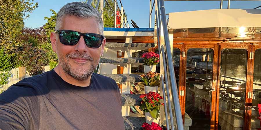  Man stood on the sunny open deck of the barge with colourful flowers in the background and wooden doors. 