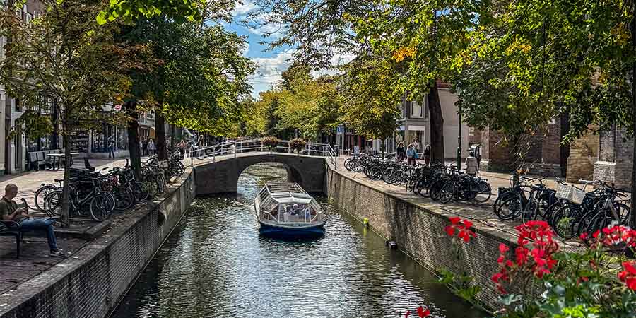 Canal boat passing beneath a stone bridge in Delft, with bicycles lining the canal and trees casting shade along the waterfront. 