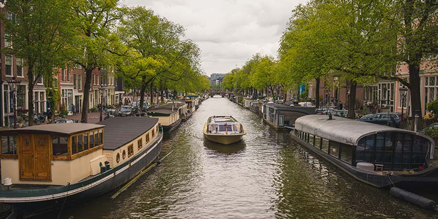 Boat travelling along an Amsterdam canal lined with houseboats, historic buildings, and leafy trees.