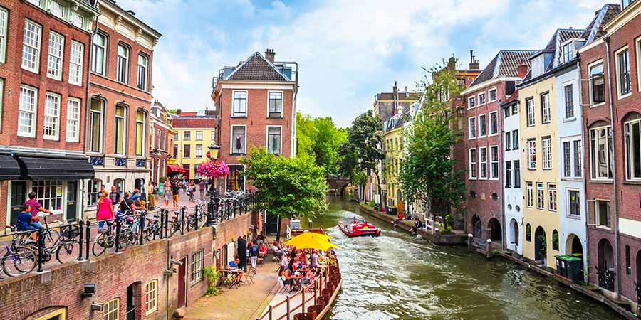 Historic buildings line a canal in Utrecht, with cyclists, cafés, and boats creating a lively waterfront scene.