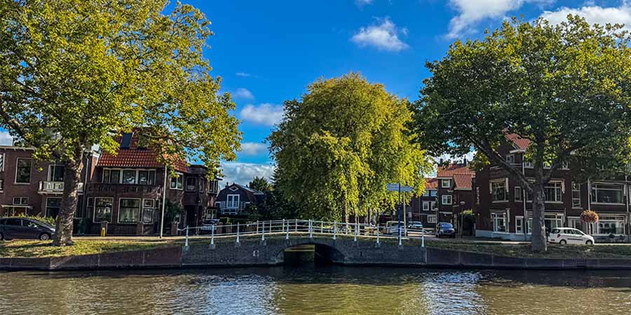Tree-lined canal with a small white bridge in Delft, surrounded by traditional brick houses under a bright blue sky. 